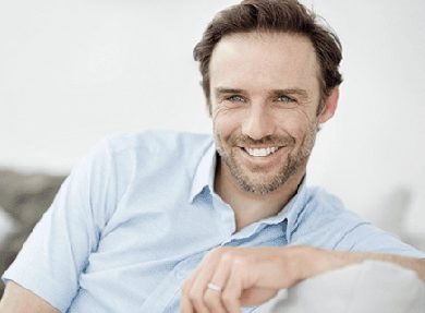 Head-and-shoulders photo of a brunette, middle-aged man sitting on a light-colored sofa and smiling, for information on cater-to-cowards dentistry from Hoffman Estates dentist Dr. William Becker.